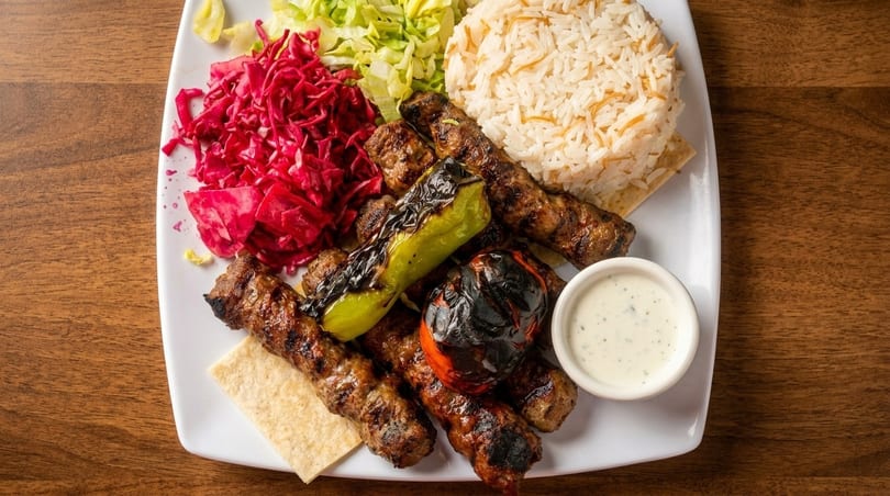 Grilled meat skewers with rice, red cabbage, green cabbage, grilled pepper, tomato, and sauce.